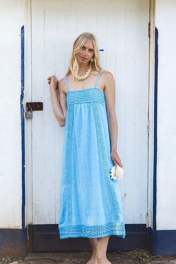 A woman stands barefoot in front of a white wooden door wearing a light blue sleeveless maxi dress with embroidered details. She is holding a small white seashell and wearing a shell necklace. Location Fingal Head, NSW.