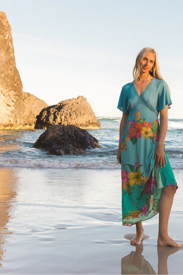 A woman stands barefoot on a sandy beach at the water's edge, wearing a teal short-sleeve maxi dress with a colorful floral print. Large rock formations and the ocean are visible in the background under a clear sky.