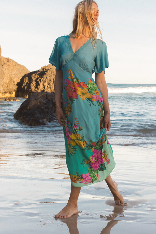 A woman wearing a teal short-sleeve midi dress with a colorful tropical floral print walks barefoot on a sandy beach with ocean waves and rocks in the background.