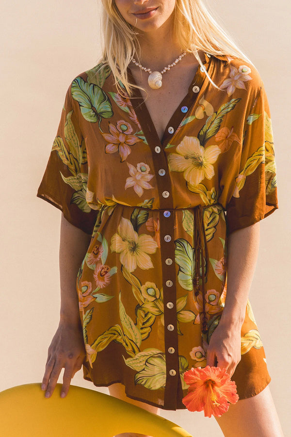 A woman wearing a brown short-sleeve button-front romper with a tropical floral print in green, yellow, and orange, holding a bright yellow beach chair against a warm beige background.