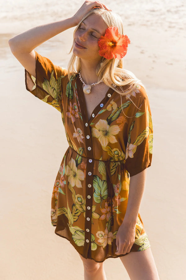 A woman on a sandy beach wearing a brown short-sleeve button-front dress with a colorful tropical floral print. She has a large orange hibiscus flower tucked into her hair and is wearing a pearl necklace. Location Dreamtime Beach, Fingal Head, NSW.