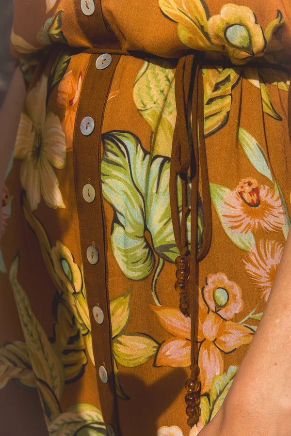 Close-up of a brown floral dress with a vertical button placket and a matching drawstring belt, featuring large green and peach flowers on a warm brown background. 