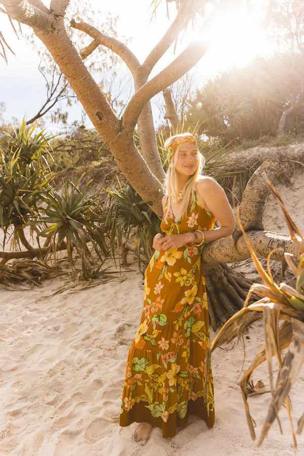 A woman stands on a sandy beach wearing a long sleeveless maxi dress with a yellow, orange, and green floral print. She is posed under the branches of a tree with bright sunlight shining through the background. Location Dreamtime Beach, Fingal Head, NSW.