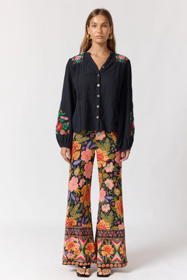 Woman wearing a black blouse with floral embroidery and colorful patterned pants on a white background