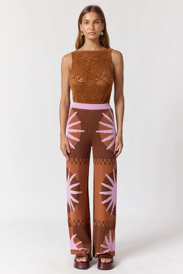 A woman modeling a sleeveless jumpsuit with a brown textured lace bodice, a light pink waistband, and wide legs featuring a brown and pink sunburst pattern, set against a plain light gray background.