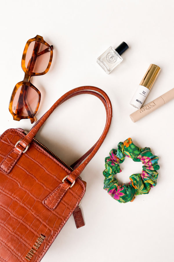 A styled flat-lay featuring a brown croc-embossed leather handbag as the main product, surrounded by accessories including tortoiseshell sunglasses, a small glass bottle, a white cosmetic tube with a gold cap, a beige lip product, and a green and pink patterned scrunchie on a white background.