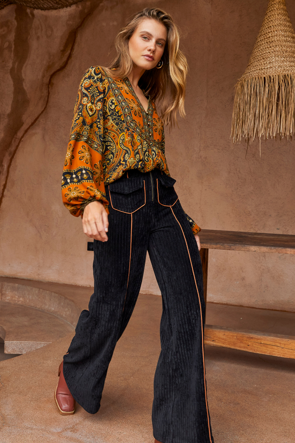 Woman wearing a patterned blouse and black pants in a warm-toned interior setting
