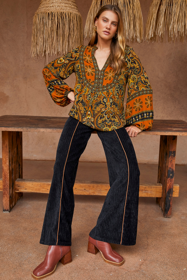 Woman wearing a patterned blouse and jeans, standing against a rustic wooden bench.