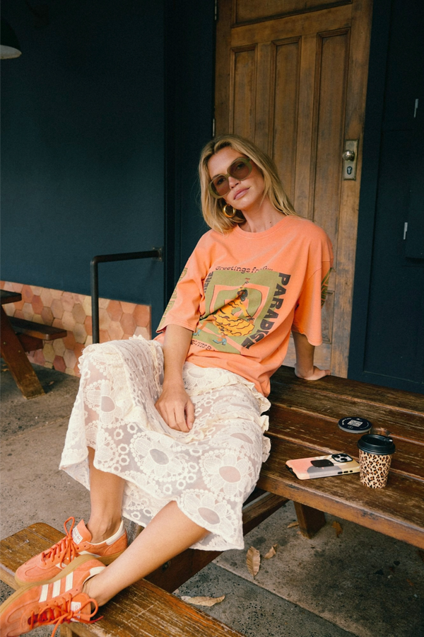 A woman sits on a wooden bench wearing a peach graphic t-shirt, a white lace midi skirt, and orange sneakers. A coffee cup and a phone are on the bench next to her.