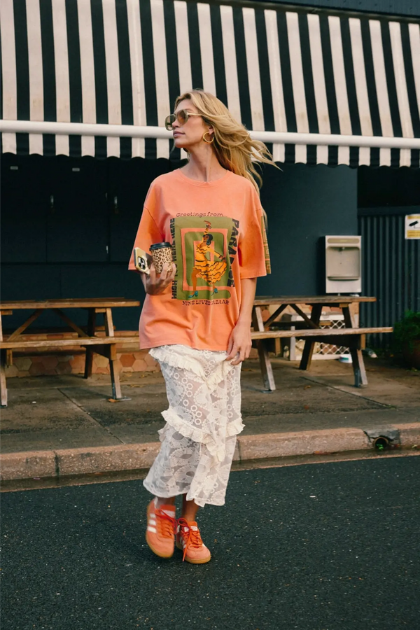 A woman standing outside a café with a black and white striped awning, wearing an oversized peach graphic T-shirt, a cream lace ruffle tiered maxi skirt and orange sneakers.