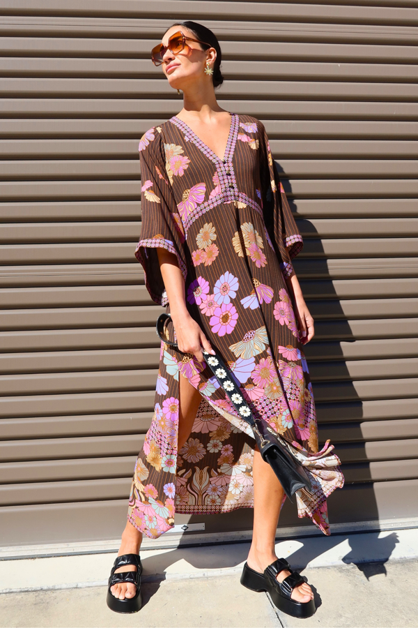 A woman wearing a brown bohemian floral kaftan dress with pink, purple, and orange flowers, holding a black and white patterned clutch bag, standing in front of a brown corrugated metal garage door.